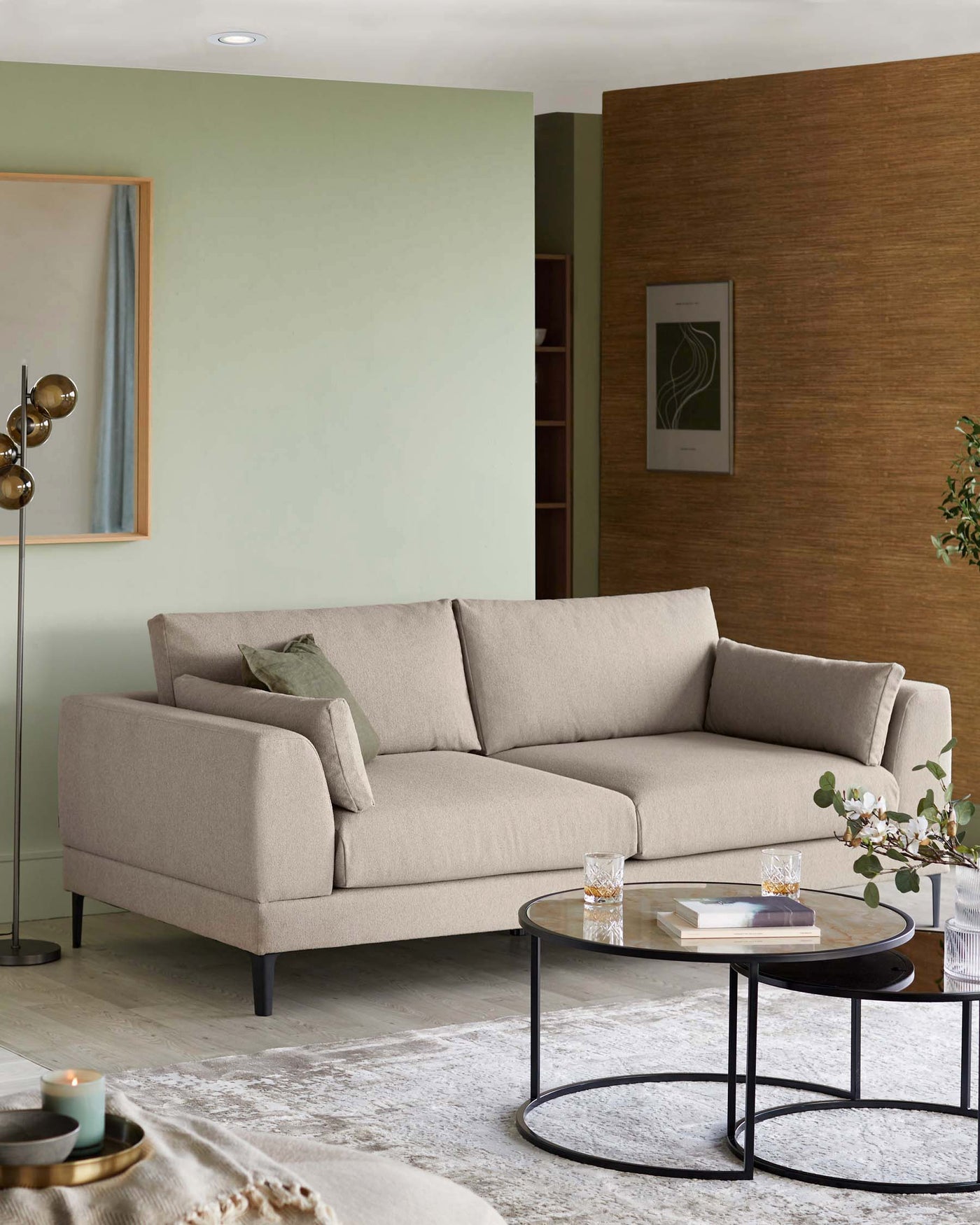 A contemporary beige sectional sofa with clean lines and dark tapered legs. In front of it is a round coffee table featuring a black metal frame with a glass top.