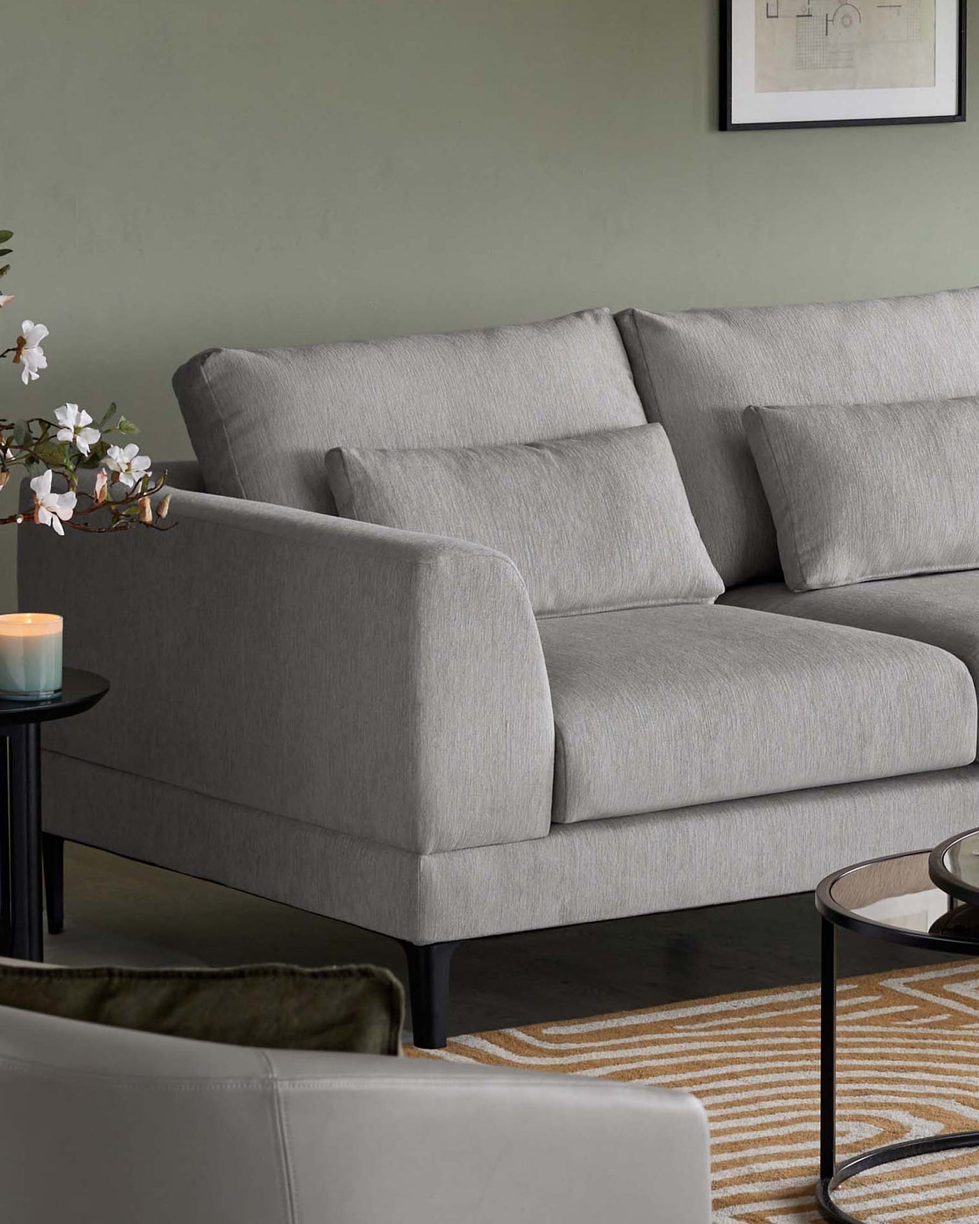Elegant modern three-seater sofa with a sleek profile, upholstered in light grey textured fabric, featuring plush back cushions and a single seat cushion, complemented by slim, dark tapered wooden legs. Beside the sofa is a minimalist round side table with a black top and matching slender legs, paired with a small decorative candle. The setting includes a muted green wall and a patterned rug with earthy tones partially visible, enhancing the contemporary aesthetic of the space.