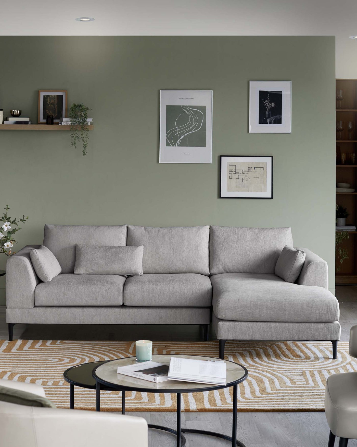 Modern minimalist living room featuring a light grey upholstered L-shaped sectional sofa with five back cushions and a chaise lounge end. In front of the sofa, there is a round black coffee table with a glass top. The furniture is placed on a beige area rug with geometric patterns.