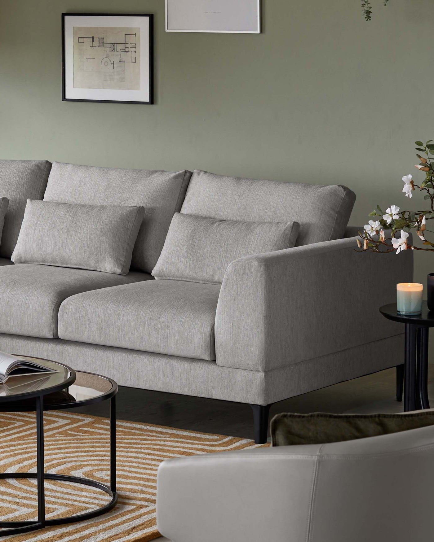 Modern three-seater sofa with grey fabric upholstery and streamlined silhouette, featuring clean lines and a minimalist design. Accompanied by a sleek round side table with a black frame and glass top, placed on a geometric-patterned area rug with cream and mustard hues.
