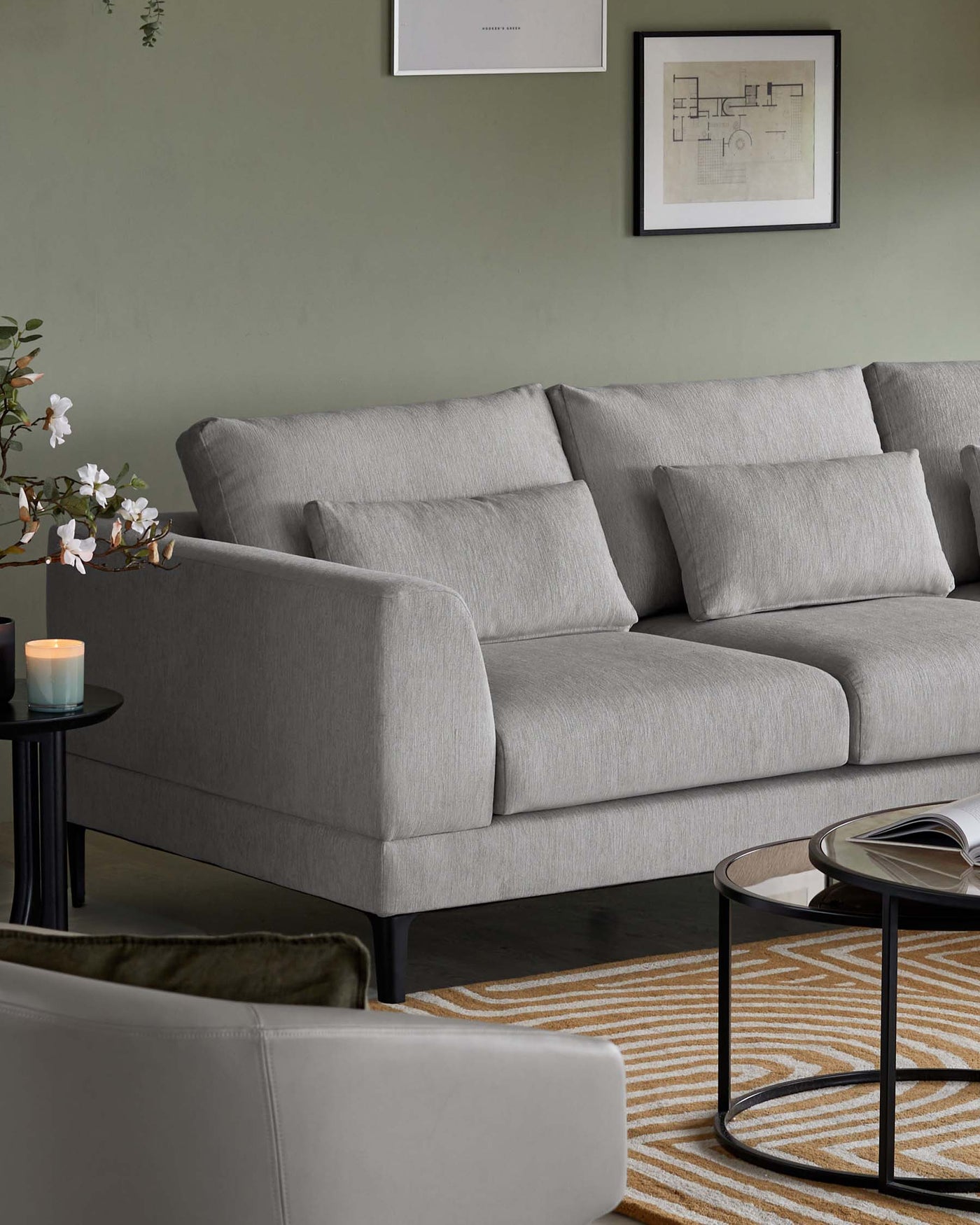 A contemporary three-seater sofa with a light grey fabric upholstery and plush back cushions. The sofa features a simple, streamlined silhouette with rounded armrests and black tapered legs. In front of the sofa, there is a round black coffee table with a clear glass top and a lower shelf with a circular pattern design. The furniture is set upon a textured yellow and white striped area rug, adding a subtle touch of colour to the space.