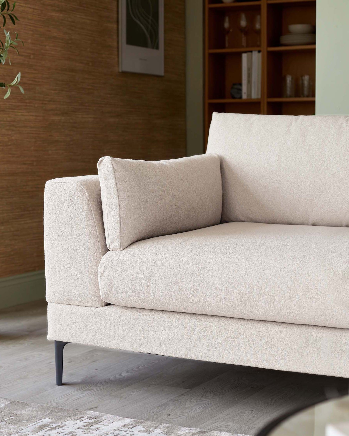 Modern light beige three-seater sofa with a minimalist design, featuring clean lines, a single seat cushion, two back cushions, and slender metal legs. A wooden bookshelf with compartments, some filled with dinnerware and decorative items, is visible in the background.