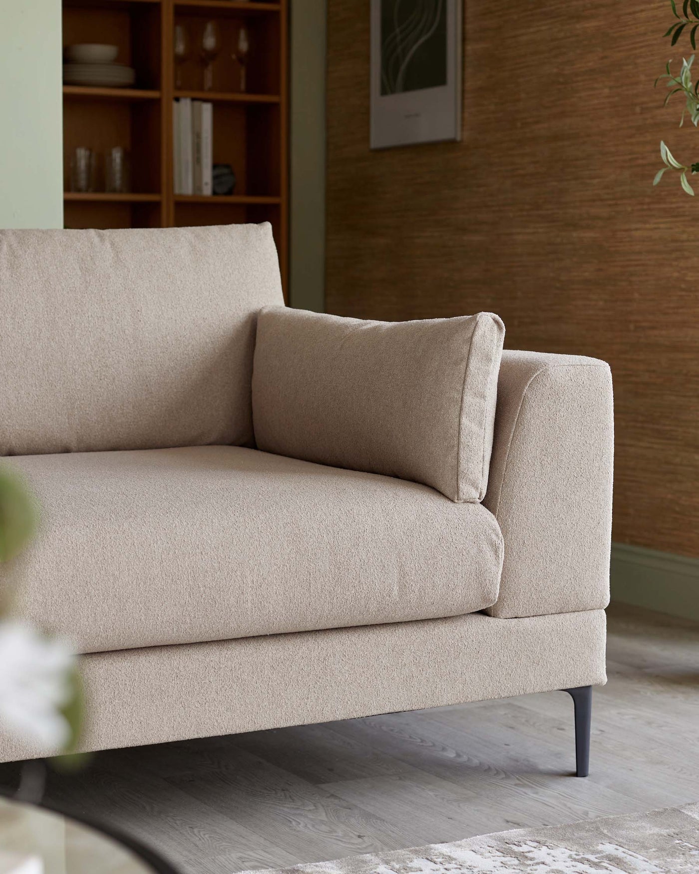 A contemporary beige sofa with a minimalistic design, featuring a smooth fabric finish, plush back cushion, and a single side cushion. The couch has a clean-lined silhouette and stands on slender, tapered dark legs, situated in a room with a wooden cabinet and artwork in the background.