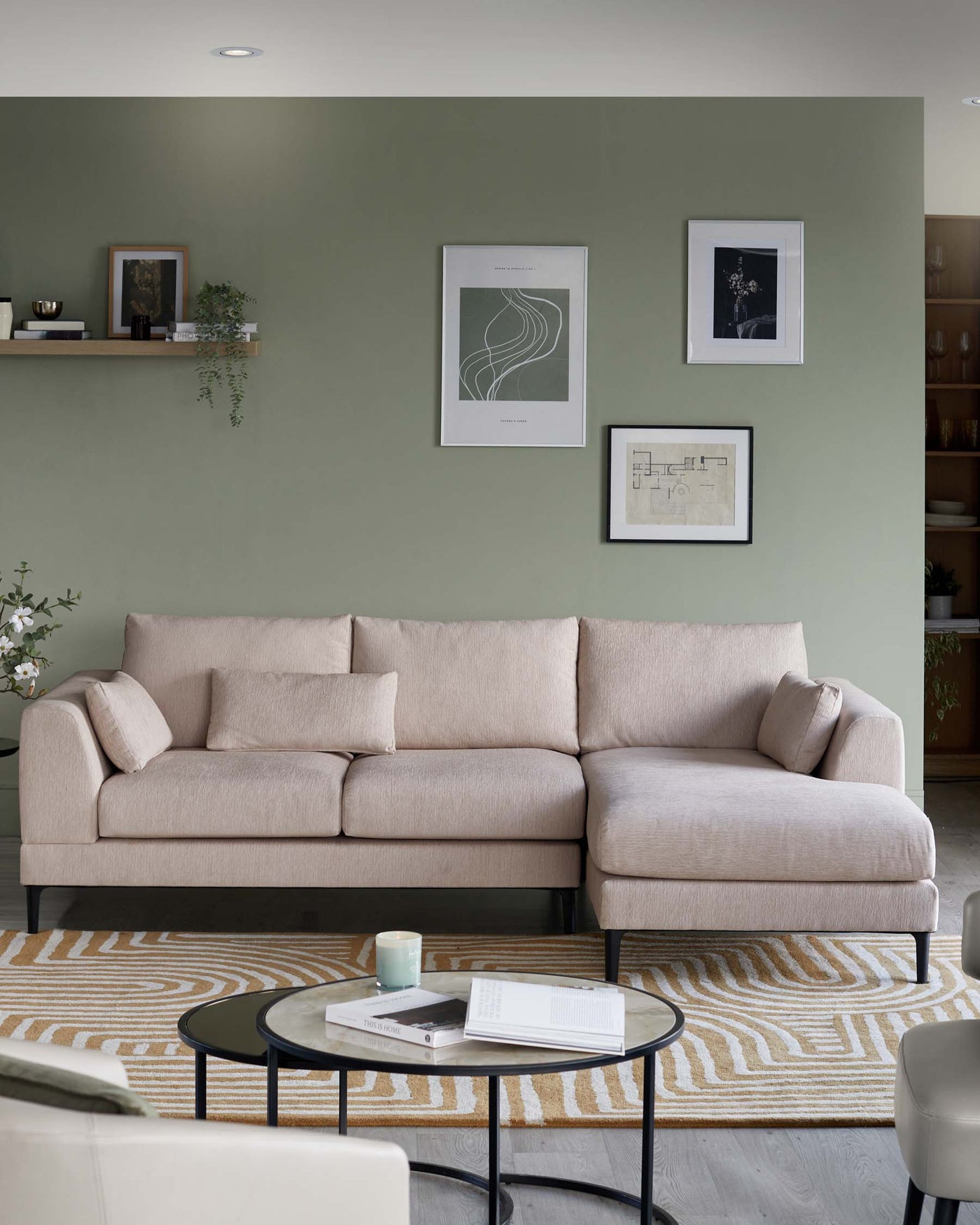 A contemporary beige sofa with a chaise lounge on the right side, upholstered in a textured fabric, featuring a low backrest with three loose back cushions, and an angled armrest on the left. The sofa legs are slim and black, providing a subtle contrast. In front of the sofa stands a round, black metal coffee table with a glossy tabletop, upon which rest a candle, two books, and a mug. The setting is complemented by a beige and yellow patterned area rug beneath the table.