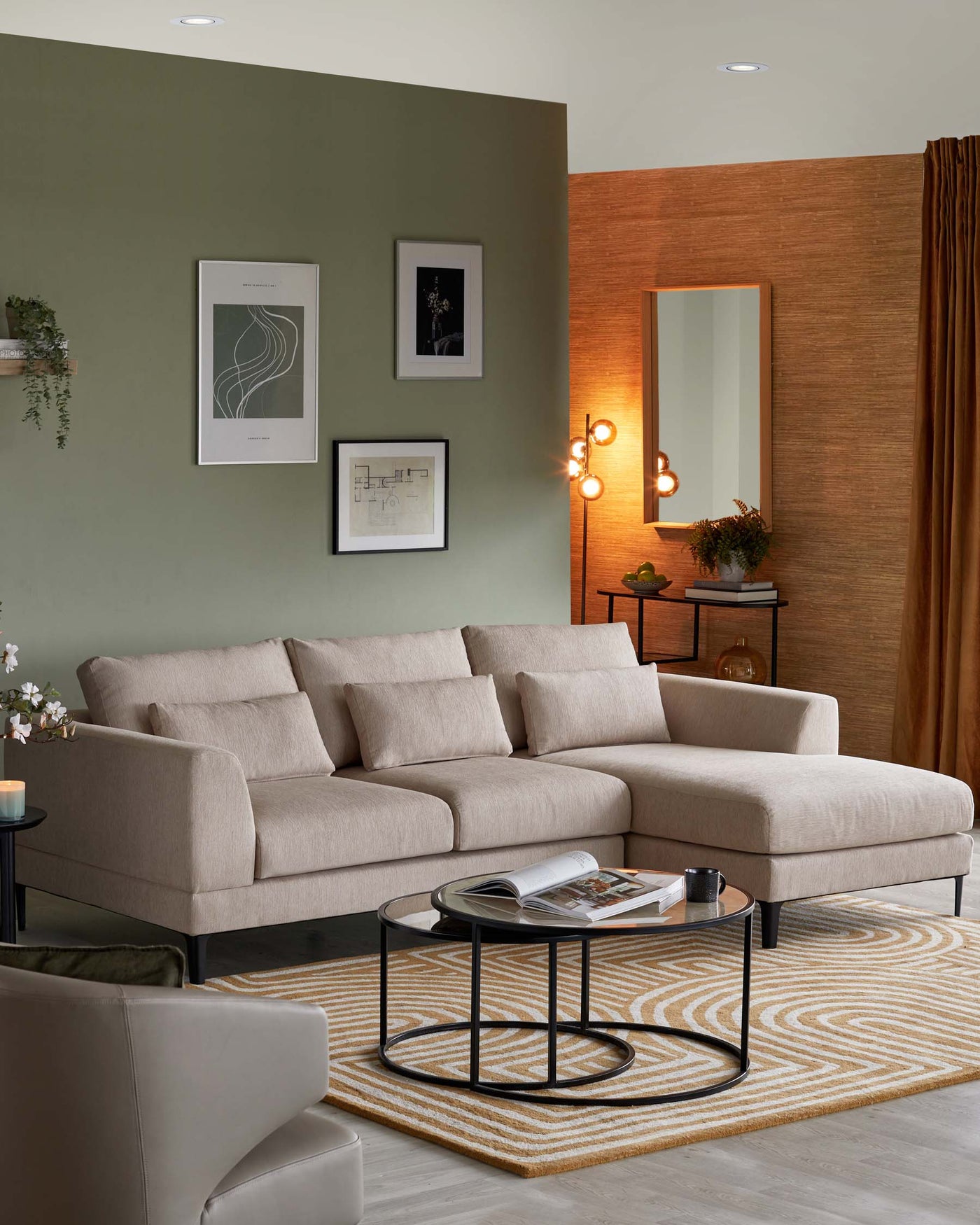 An elegant, contemporary living room setup featuring a beige L-shaped sectional sofa with plush back cushions and a chaise lounge end. A set of round nesting coffee tables with black metal frames and glass tops sits on a beige and yellow patterned area rug. An accent side table with a dark top and matching black legs is placed beside a minimalist wooden console with a large mirror above it. The room conveys a warm and inviting atmosphere with tasteful decor.