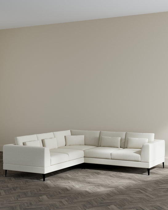 Modern L-shaped sectional sofa with chaise in a light cream upholstery and dark wooden legs, set against a minimalistic interior with herringbone hardwood flooring.