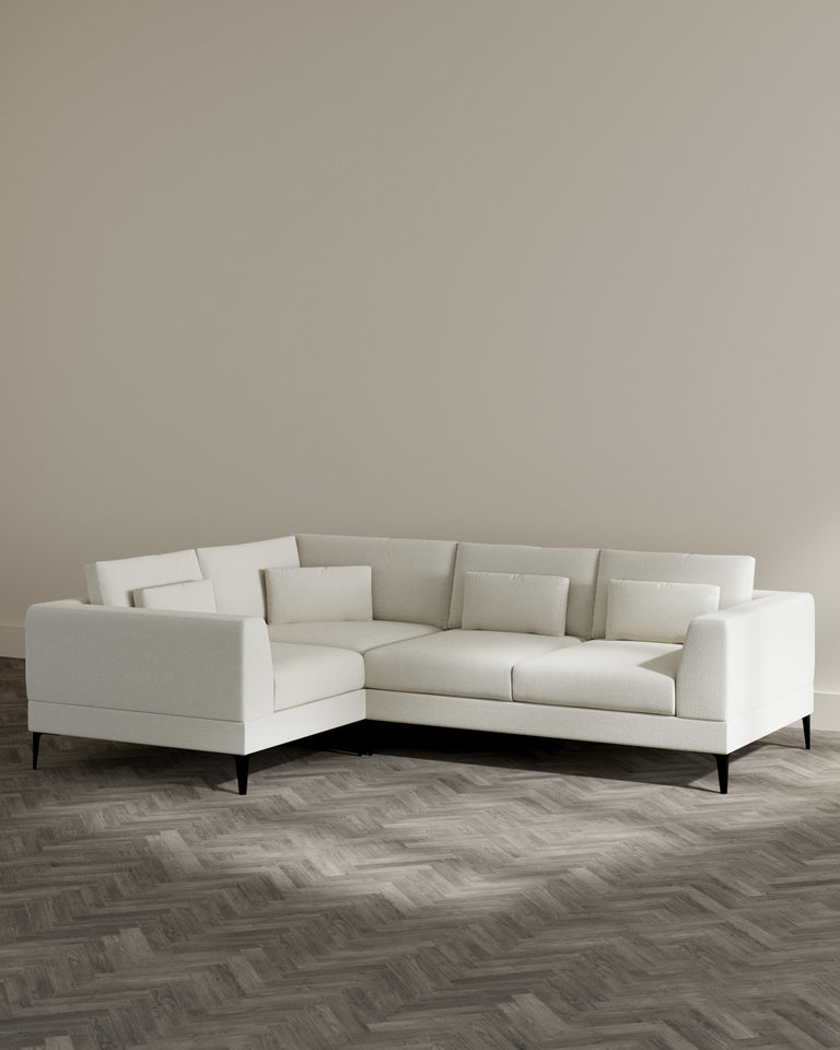 Modern L-shaped sectional sofa in a light beige upholstery with clean lines, a low back, and black tapered legs, set in a room with herringbone wood flooring.