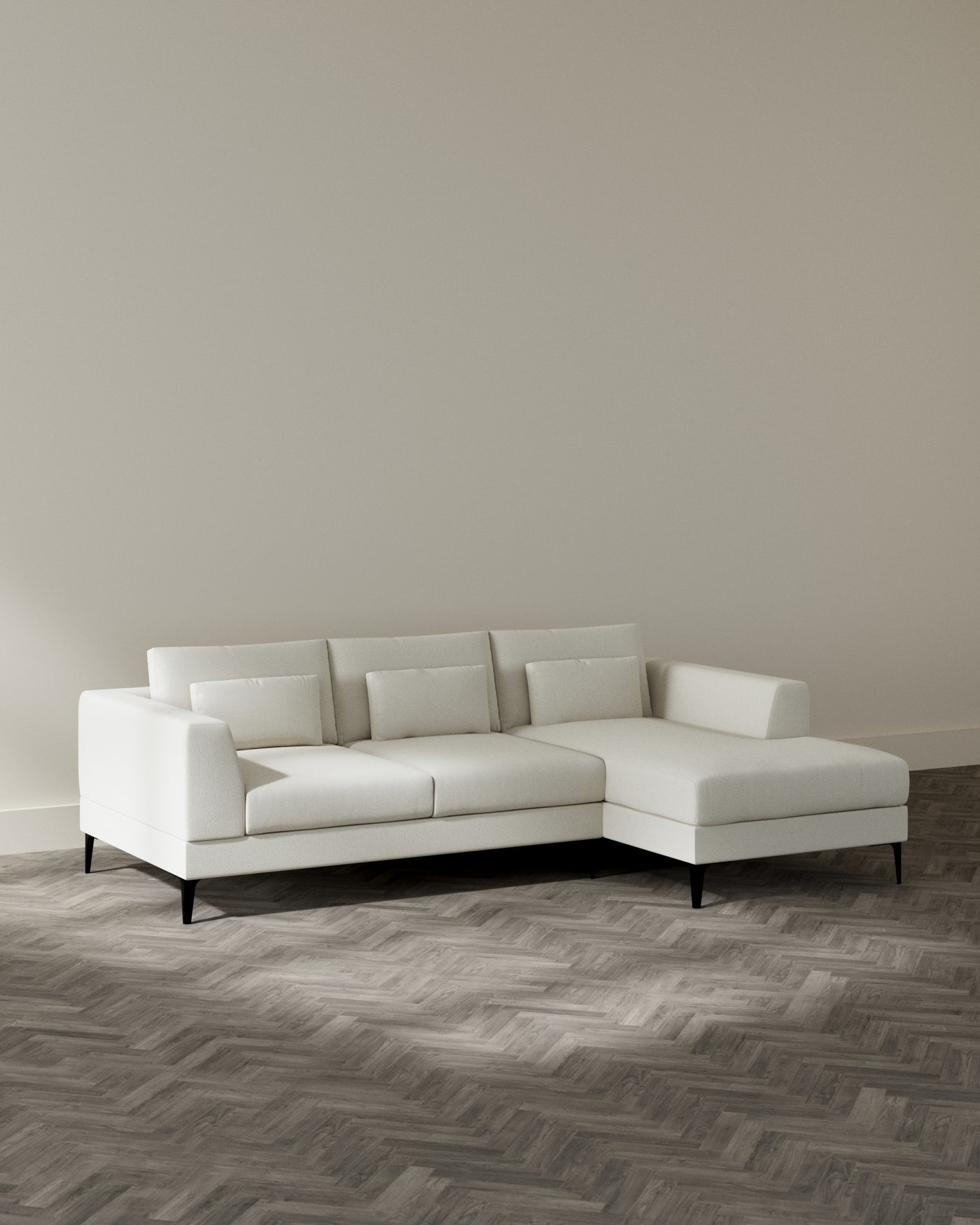 Modern L-shaped sectional sofa with a left-hand chaise lounge, upholstered in a light cream fabric, featuring clean lines, plush back cushions, and tapered black legs set against a neutral-toned wall and herringbone patterned wooden flooring.