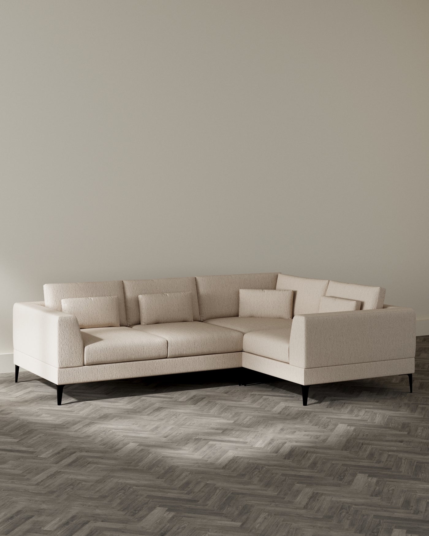 An L-shaped sectional sofa with a modern design, featuring a textured light beige fabric upholstery and clean, straight lines. The sofa has plush back cushions and a uniform seat cushion, elegantly elevated on slender black legs that provide a subtle contrast to the light upholstery. The sofa sits on a herringbone-patterned wooden floor, and the room has a neutral colour palette.