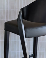 Black leather bar stool with curved backrest and wooden armrest, shown at an angle against a white tiled wall.