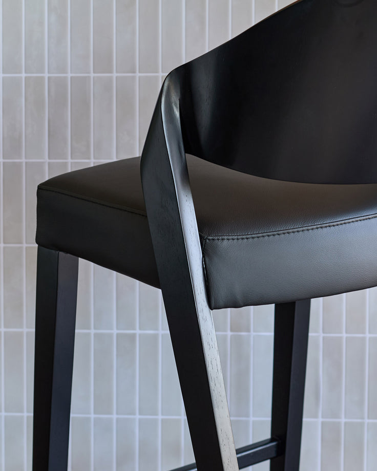 Black leather bar stool with curved backrest and wooden armrest, shown at an angle against a white tiled wall.