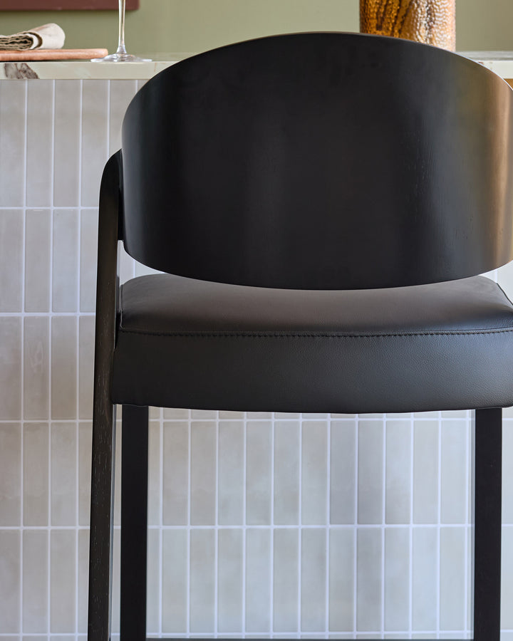 Enzo black leather bar stool with curved backrest and dark wooden legs, side view detail.