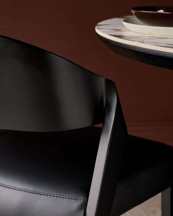 Enzo dining chair in black leather, side view showing curved backrest and metal frame detail against brown background.