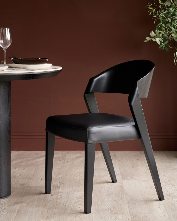 Enzo dining chair in black leather with dark wood frame, shown at round table setting with place setting and greenery.
