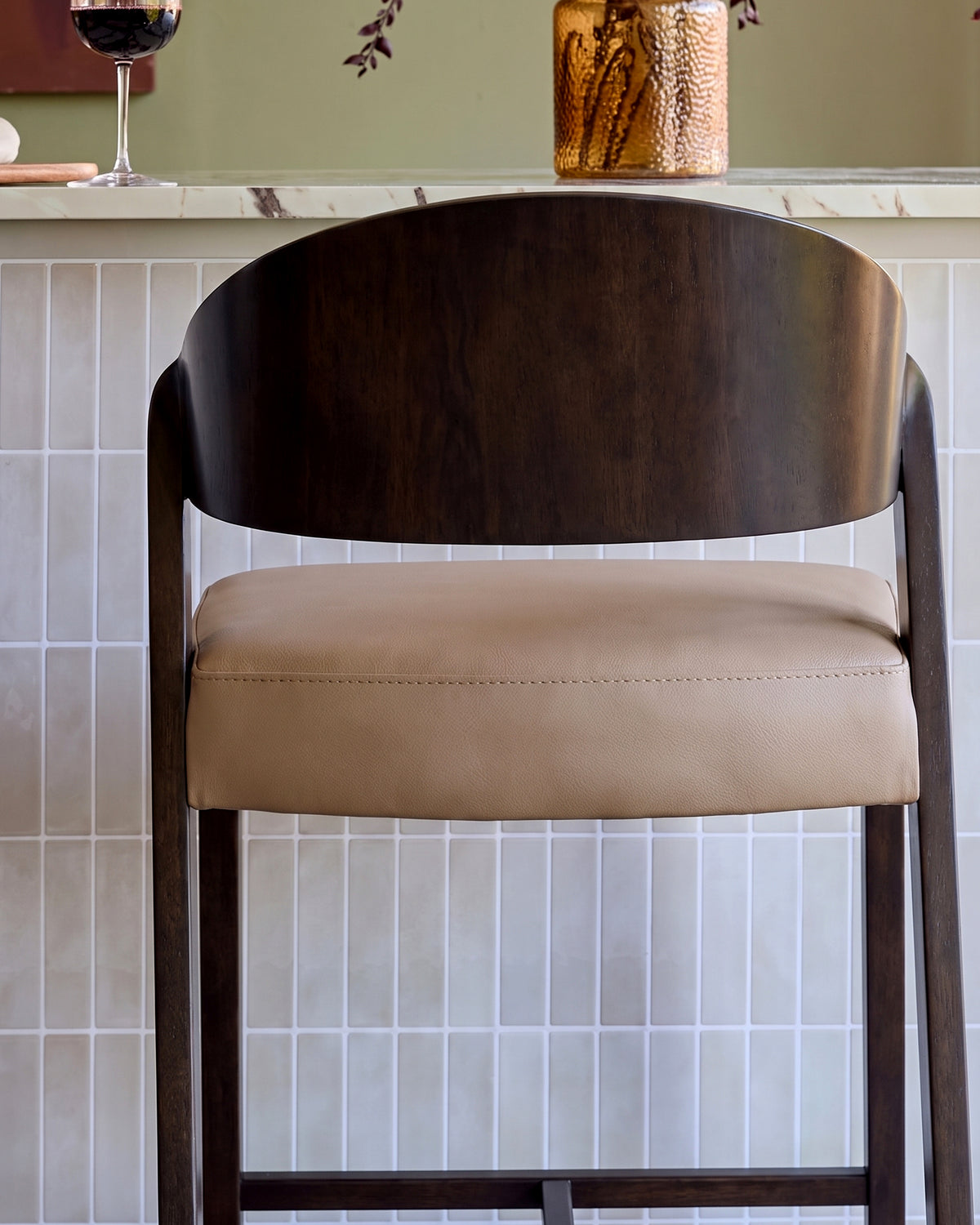 Enzo bar stool in dark wood with mocha leather seat, shown front view against white subway tile wall.