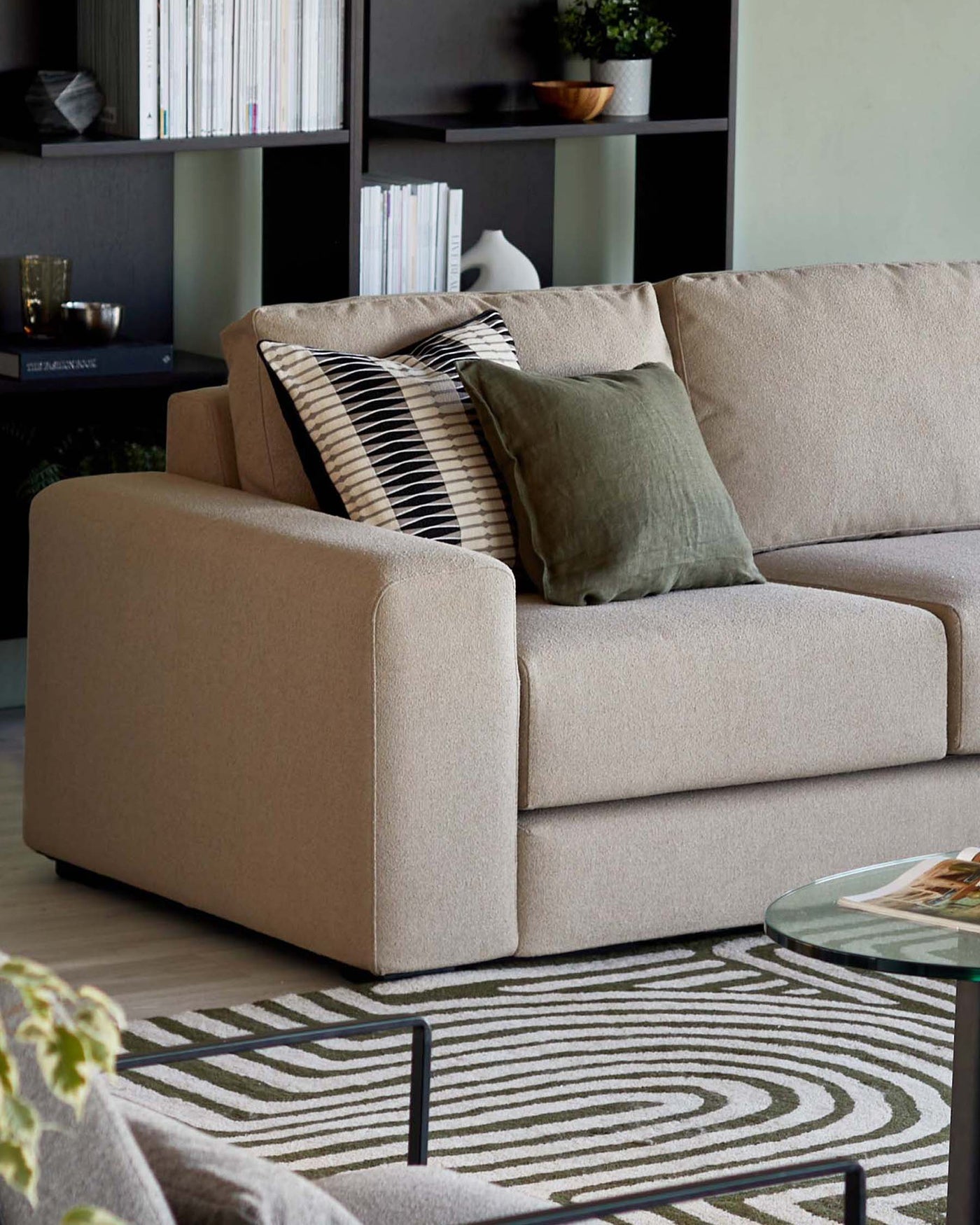 A contemporary beige fabric sofa with rounded armrests and clean lines, accented by striped and solid green throw pillows. A glass coffee table with a minimalist black metal frame is positioned in the foreground, and behind the sofa stands a dark bookcase filled with assorted books and decorative items. The furniture is set upon a patterned area rug with a geometric design.