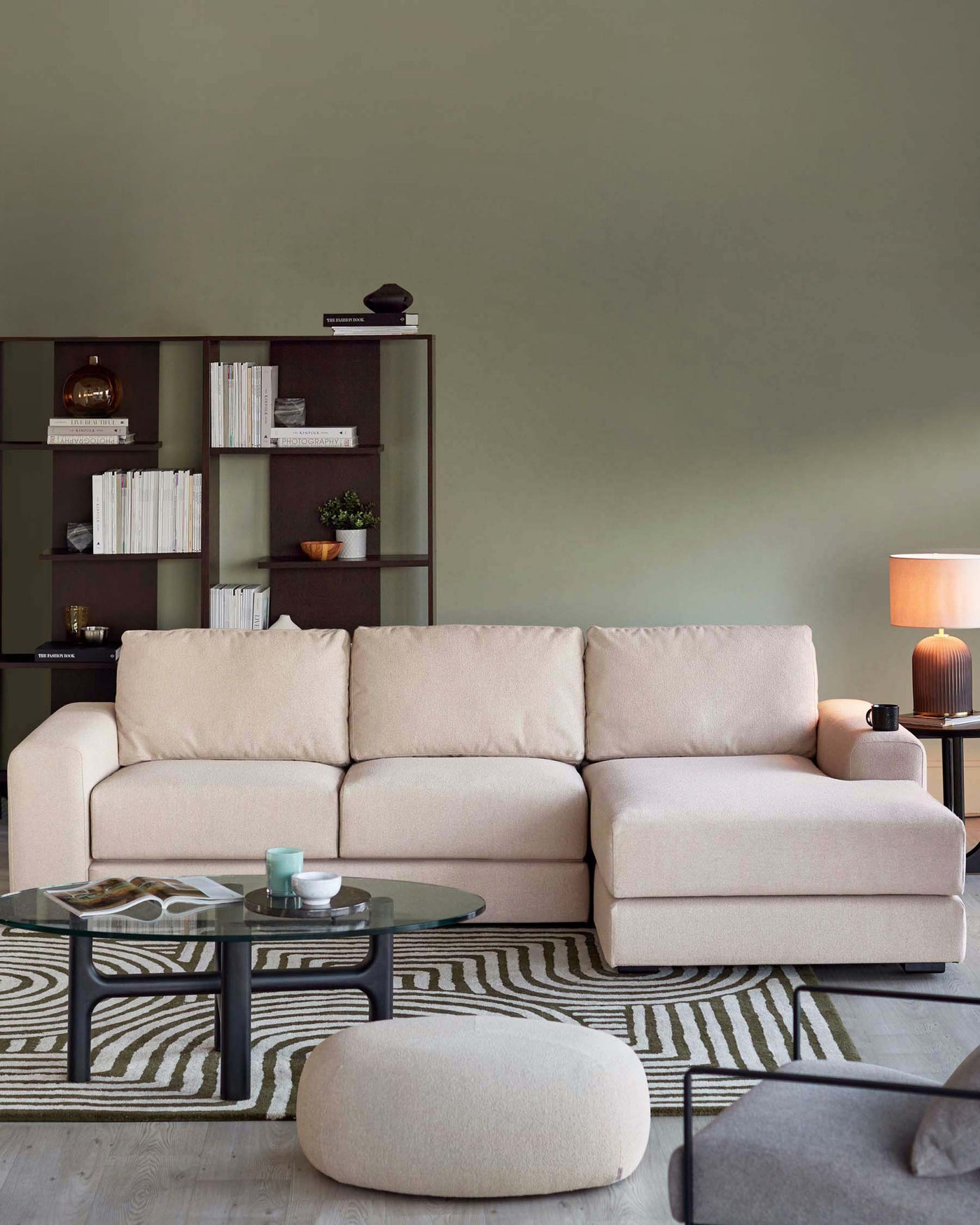 A modern living room featuring a modular beige fabric sectional sofa with clean lines and plush cushions. In front of the sofa is a round glass-top coffee table with a black metal base. To the side sits a matching beige fabric ottoman. The sofa is accessorized with small black reading lamps on either end, and the room is completed with a textured beige and grey striped area rug. Behind the sofa, there is a dark-toned wooden bookshelf filled with an assortment of books and decorative items.