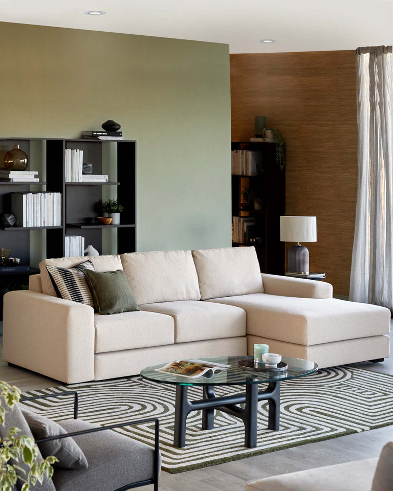 A contemporary living room setting featuring a light beige sectional sofa with plush cushions, complemented by a patterned grey and white area rug. The centrepiece is a round, glass-top coffee table with a unique black frame design. To the left, a minimalist black shelving unit displays a selection of books and decorative items.