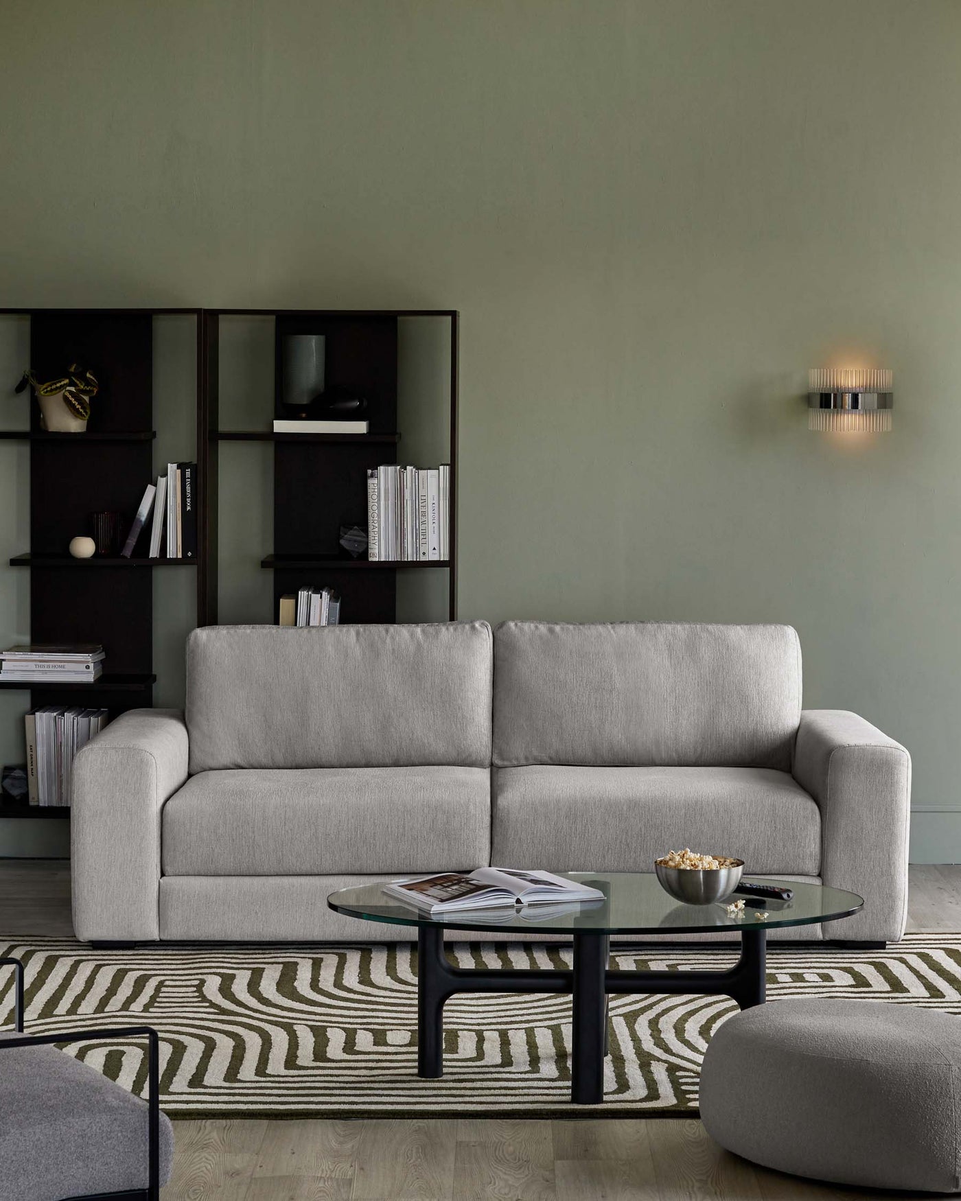 Modern living room featuring a minimalist light grey upholstered sofa with clean lines and a plush look, complemented by a sleek, oval-shaped glass-top coffee table with a black frame. An oval-shaped grey ottoman matches the sofa. The set is arranged on a patterned area rug with an undulating design, and a contemporary open shelving unit in black behind the sofa displays an assorted collection of books and decorative items.