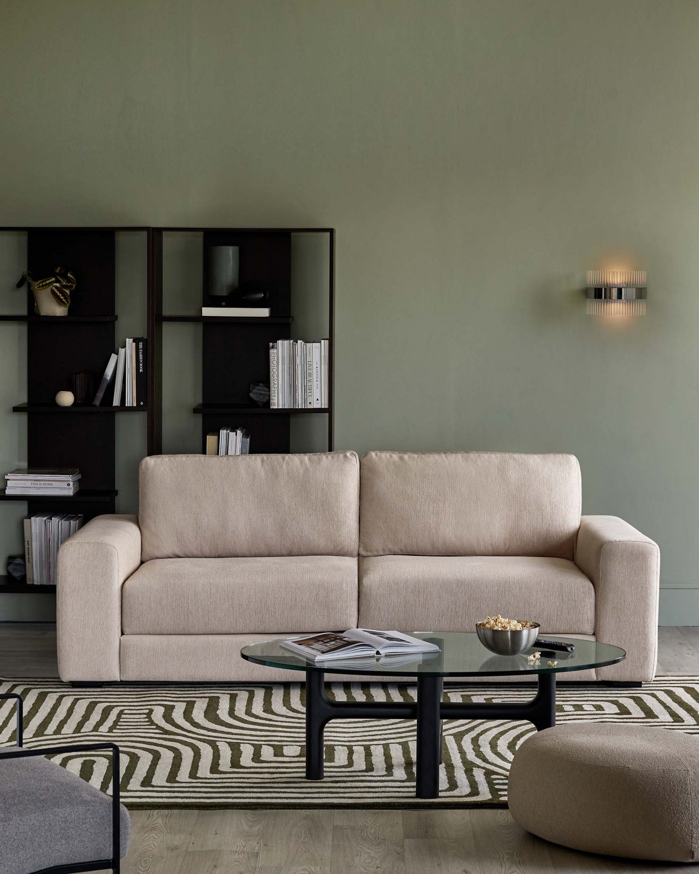 A contemporary, beige upholstered three-seater sofa with a minimalist silhouette and rounded armrests. In front of the sofa is an oval glass-top coffee table with a black frame, resting on a patterned area rug. To the left, a matching beige upholstered ottoman complements the seating. On the background wall is a dark-toned open bookshelf filled with assorted books and decorative items.