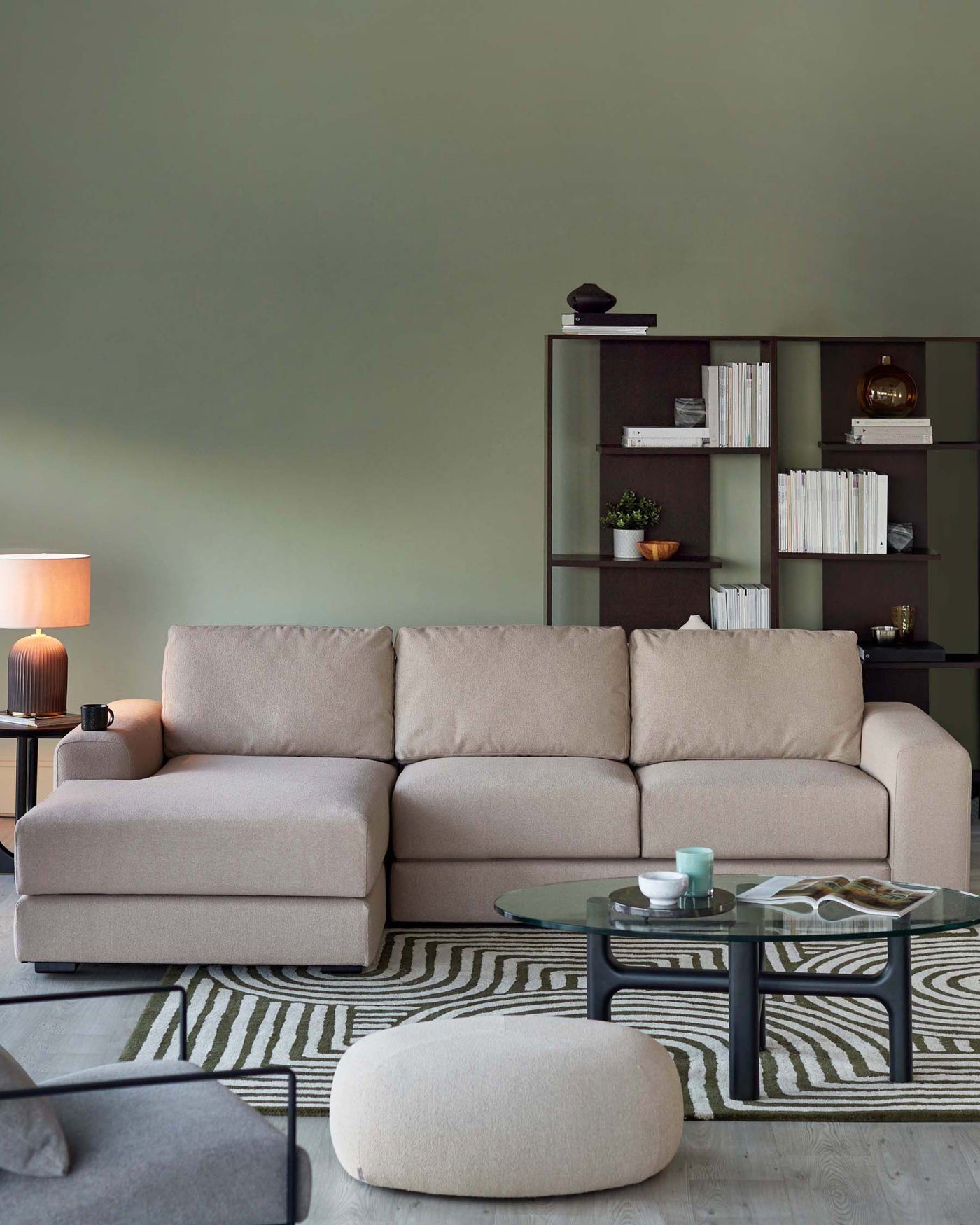 Modern living room furniture arrangement featuring a beige upholstered sectional sofa with a chaise, a round glass-top coffee table with a black frame, a dark brown shelving unit filled with books and decorative items, a beige fabric oval ottoman, a table lamp with a white shade, and a patterned area rug with green and beige tones.