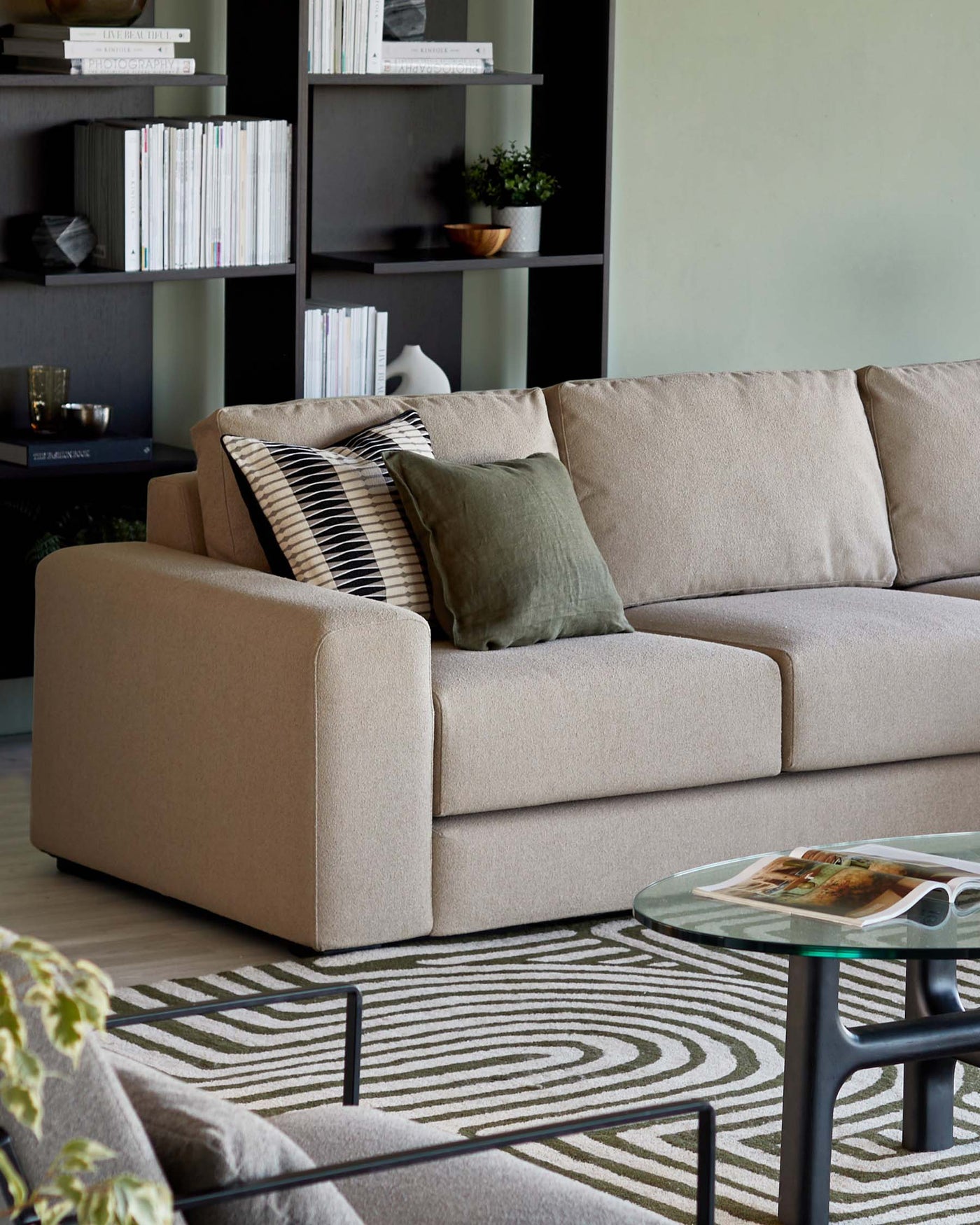 Beige upholstered three-seater sofa with a selection of decorative pillows, complemented by a glass-top coffee table with a black metal frame placed on a patterned area rug. In the background, a dark-toned bookshelf housing various books and decorative items enhances the modern living room ambiance.