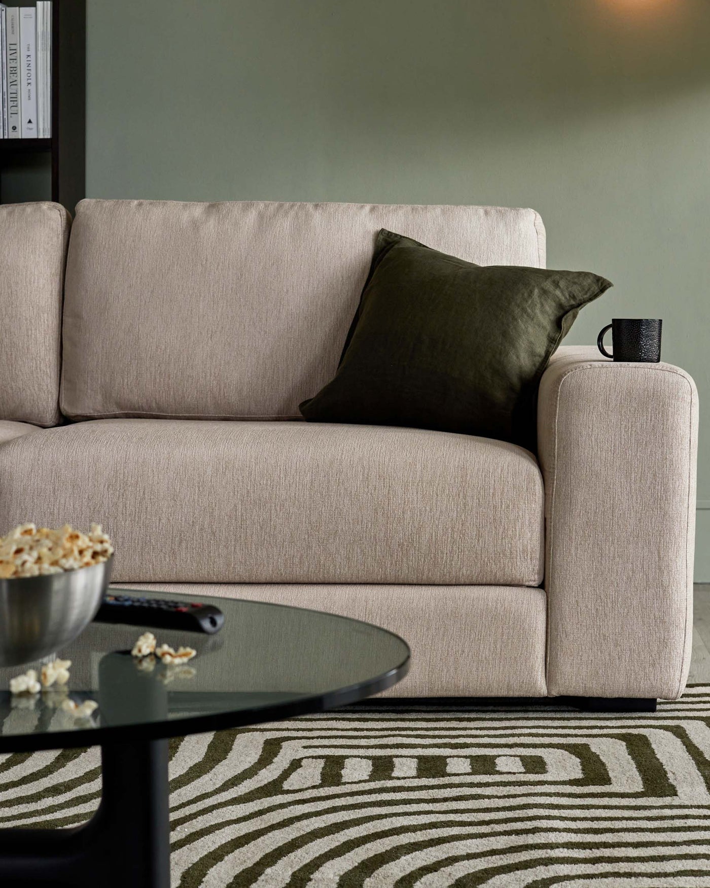 A contemporary beige fabric sofa with clean lines and a minimalist design, featuring one armrest visible on the right and a cushioned back. The sofa is adorned with a single dark olive green throw pillow and has a round armrest on one side where a black mug is placed. In the foreground, a round, smoked glass coffee table with a black frame holds a bowl of popcorn and a remote control, situated on top of a patterned area rug with geometric shapes in neutral tones.
