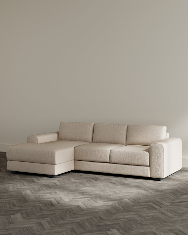 Modern beige sectional sofa with chaise lounge on left side and textured upholstery, featuring a low-profile design with clean lines and minimalistic black base, set on a herringbone wood floor against a plain light-coloured wall.