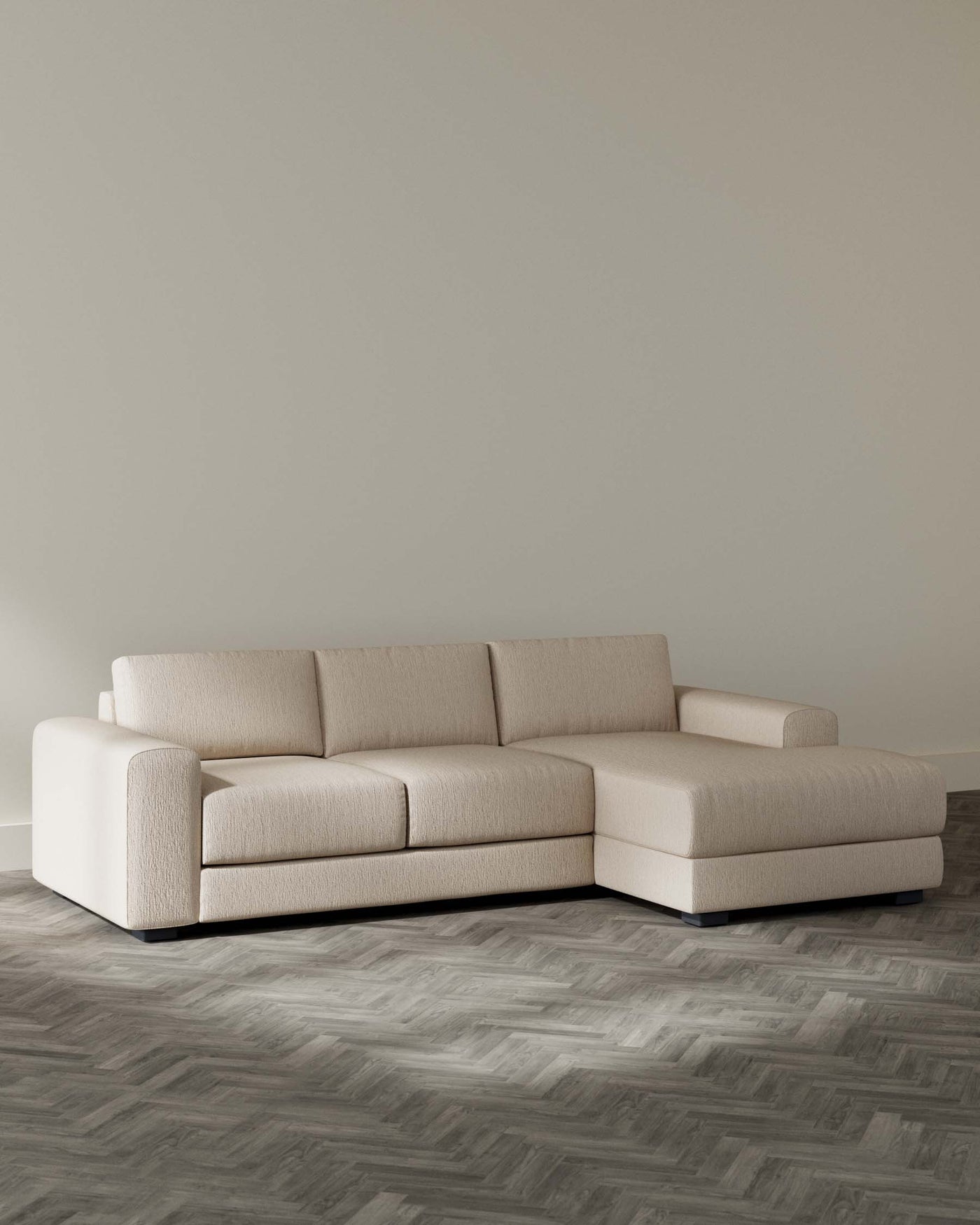 Modern beige L-shaped sectional sofa with clean lines and a plush finish, set against a neutral wall on a herringbone patterned floor.