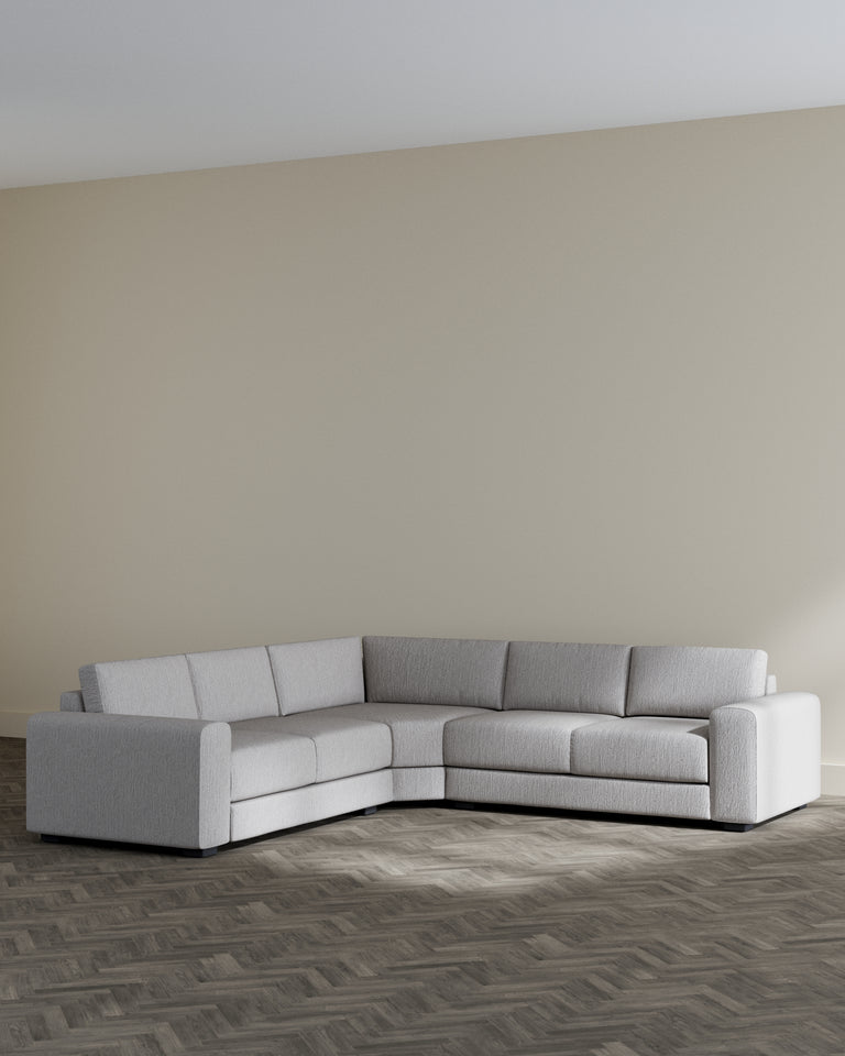 A modern light grey sectional sofa with a minimalist design, featuring clean lines and plush cushions, set on a dark herringbone patterned floor against a neutral wall.