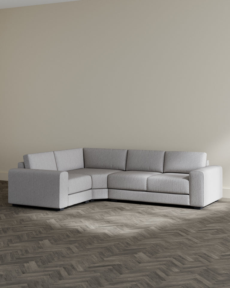 Modern light grey sectional sofa with a plush texture and a simple, contemporary design. Features clean lines, wide armrests, and a modular form that can be arranged to suit various living spaces. Set against a neutral-toned wall on a dark herringbone-patterned floor.
