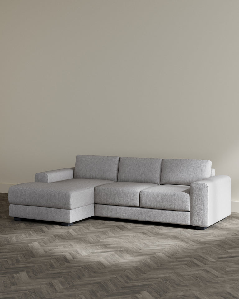 Modern L-shaped sectional sofa in light grey fabric with a chaise on the left side, featuring clean lines, a structured silhouette, and minimalistic square arms, standing on low-profile black legs. Positioned on a herringbone patterned wooden floor against a neutral wall in a minimalist setting.