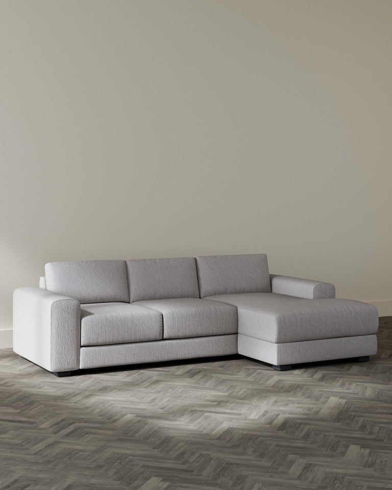 Modern light grey sectional sofa with a chaise lounge on the left side, clean lines, and plush cushions, set against a neutral wall on a herringbone patterned wooden floor.