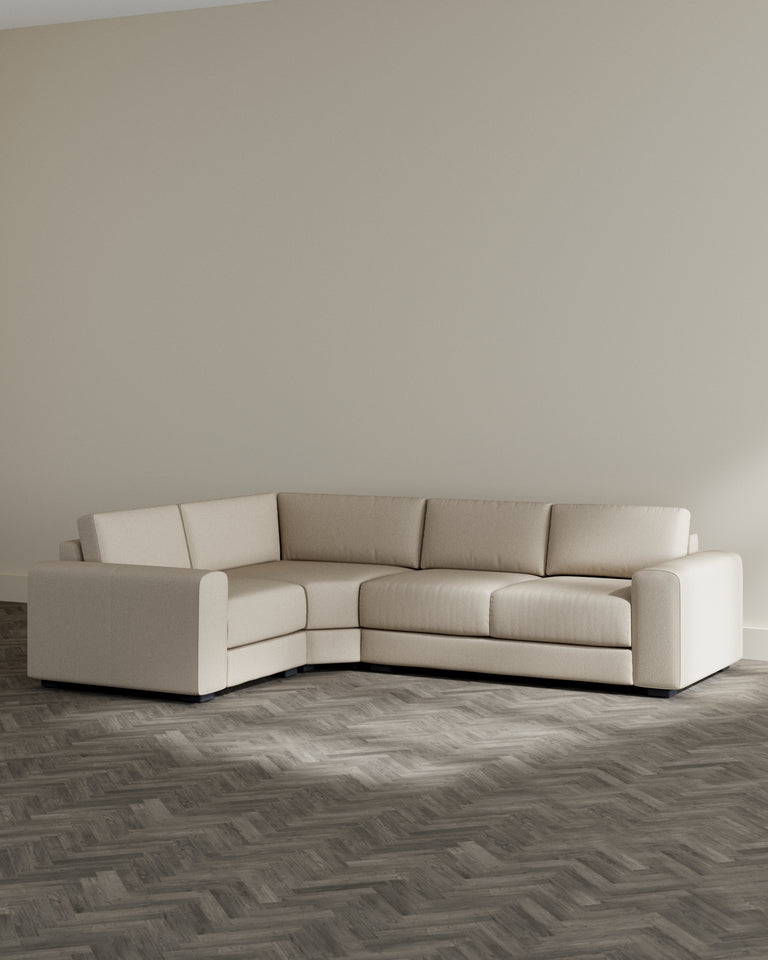 Contemporary L-shaped sectional sofa in a textured light beige fabric with cylindrical armrests and minimalistic low-profile black legs, arranged in a spacious room with light grey walls and herringbone-patterned hardwood flooring.