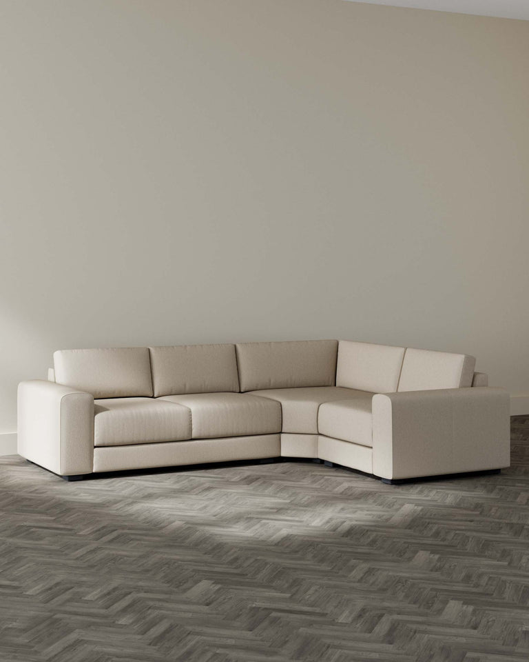 A contemporary light beige sectional sofa with a clean design, featuring a rounded corner piece and plush cushioning, set against a neutral wall on a grey herringbone patterned floor.