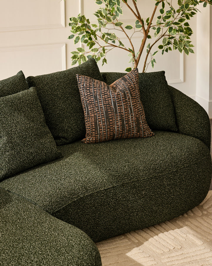 Dark green fabric left-hand corner chaise sofa with rounded arms, styled with patterned brown cushion and potted plant.