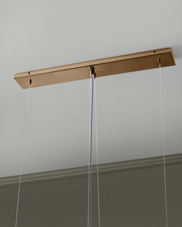 Close-up of a gold-toned ceiling light fixture with clear suspension cables. The design is sleek and modern.