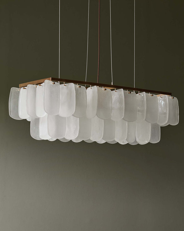 A rectangular chandelier with frosted glass panels hangs against a muted green wall, creating a stylish and modern ambiance.
