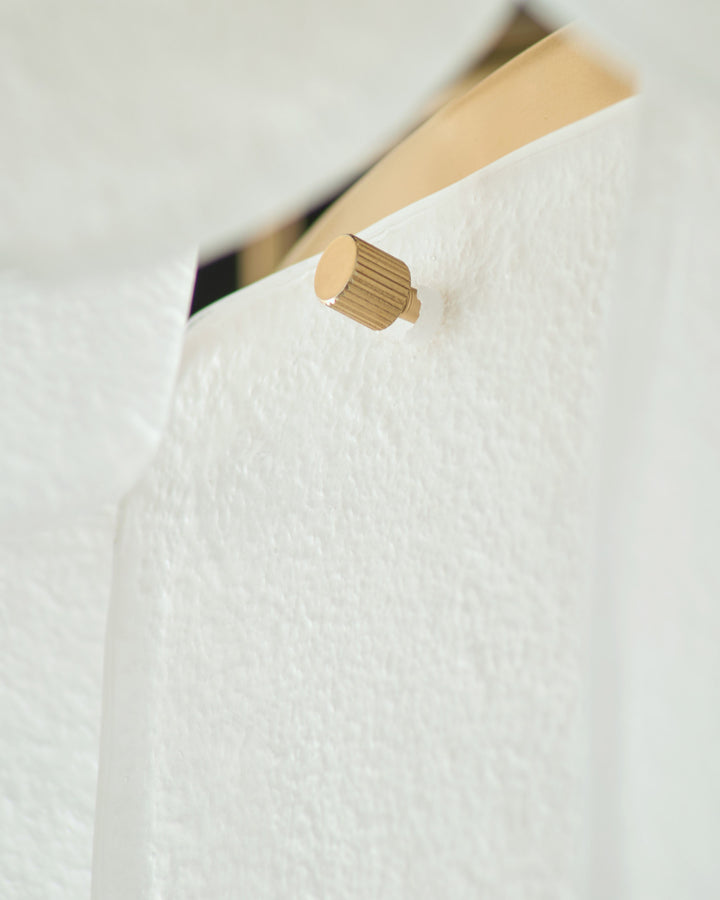 Brass cylindrical knob handle on white textured surface, angled view showing product detail.
