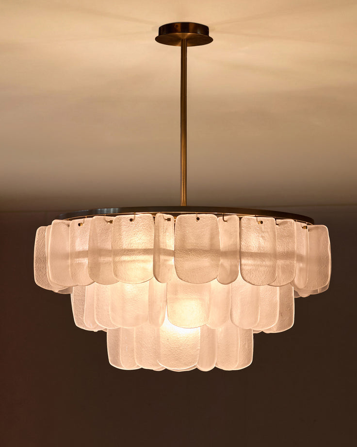 Estelle chandelier with tiered frosted glass panels in white, brass stem, suspended against warm neutral background.