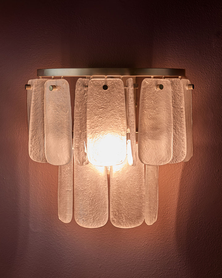 Estelle wall light with frosted glass panels in white, mounted on textured terracotta wall with warm glow from central bulb.