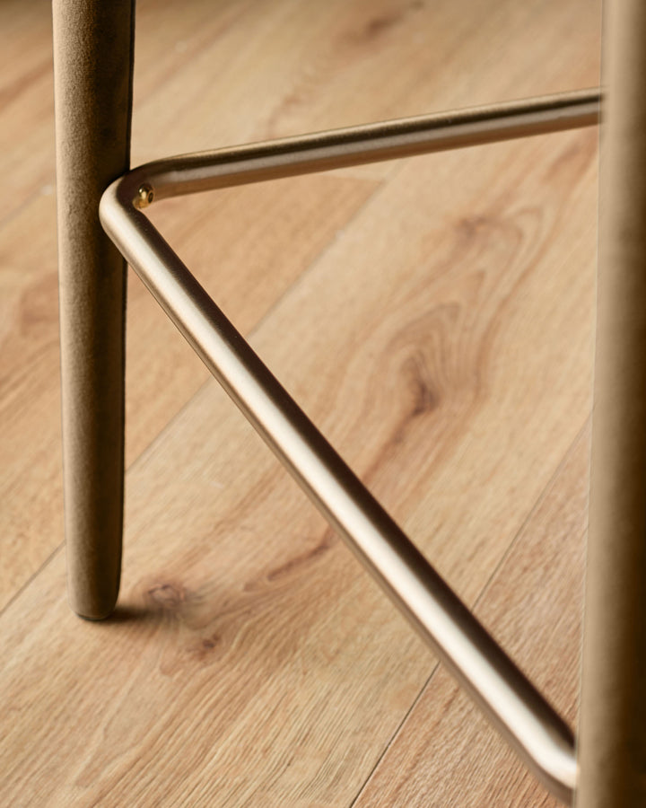 Close-up of brushed metal footrest and leg detail on caramel fabric bar stool against wooden floor.