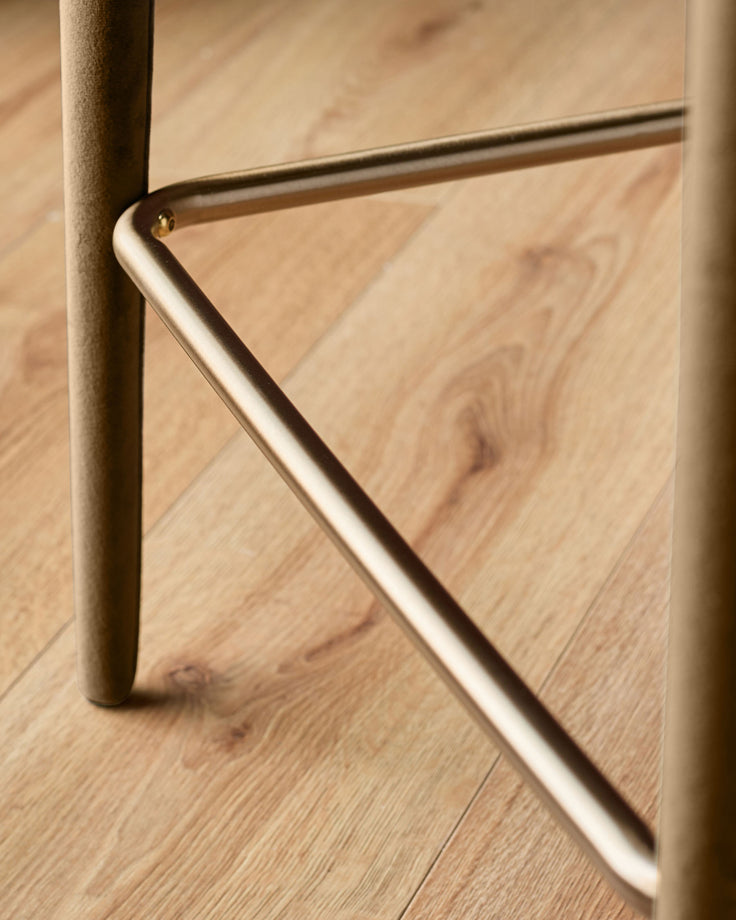 Close-up of brushed metal footrest and leg detail on caramel fabric bar stool against wooden floor.