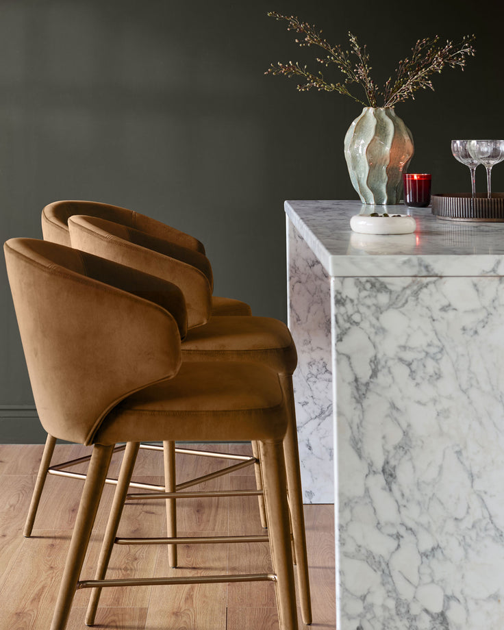 Caramel fabric bar stool with curved backrest and wooden legs, positioned at marble counter in modern interior setting.