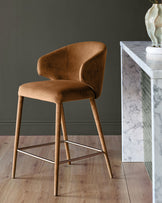 Caramel fabric bar stool with curved back, wooden legs, and metal footrest, styled in modern interior setting.