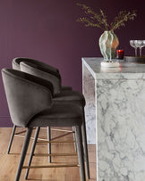 Ethan bar stool in dark chocolate velvet with curved back, metal legs, and footrest, shown at marble counter.