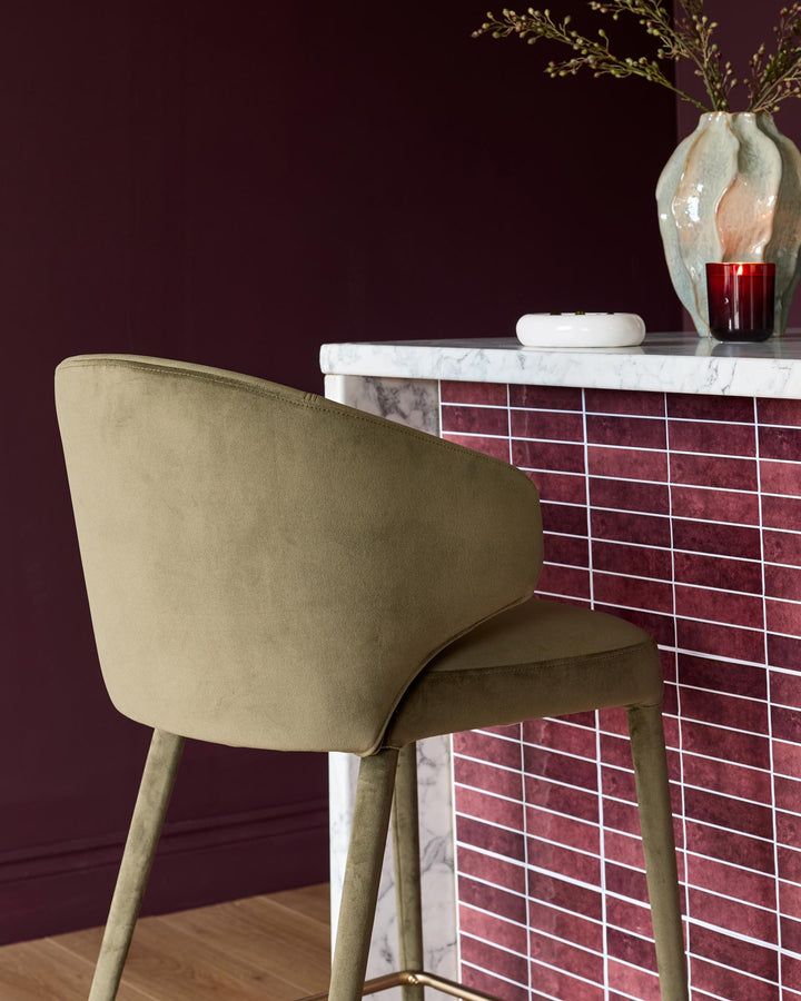 Olive green velvet bar stool with curved back and wooden legs, positioned at a burgundy tiled counter with marble top.