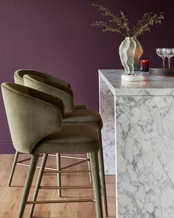 Olive green velvet bar stool with curved backrest and metal legs, shown at marble counter in styled interior.
