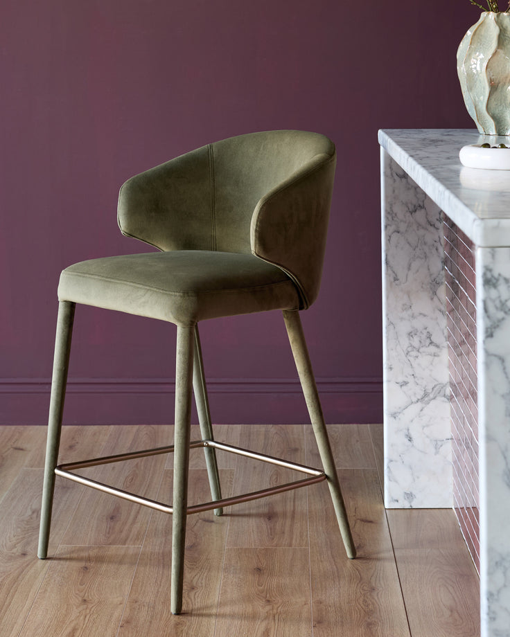 Ethan bar stool in olive green velvet with curved backrest, wooden legs, and metal footrest against purple wall.