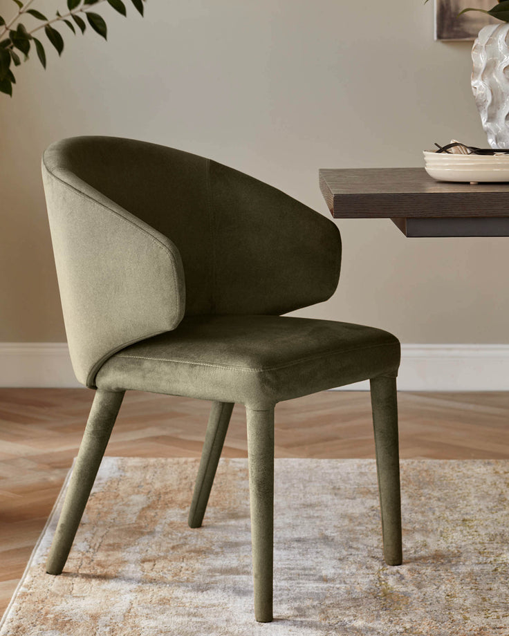 An olive green velvet chair with rounded back stands beside a dark wood table on a neutral rug.